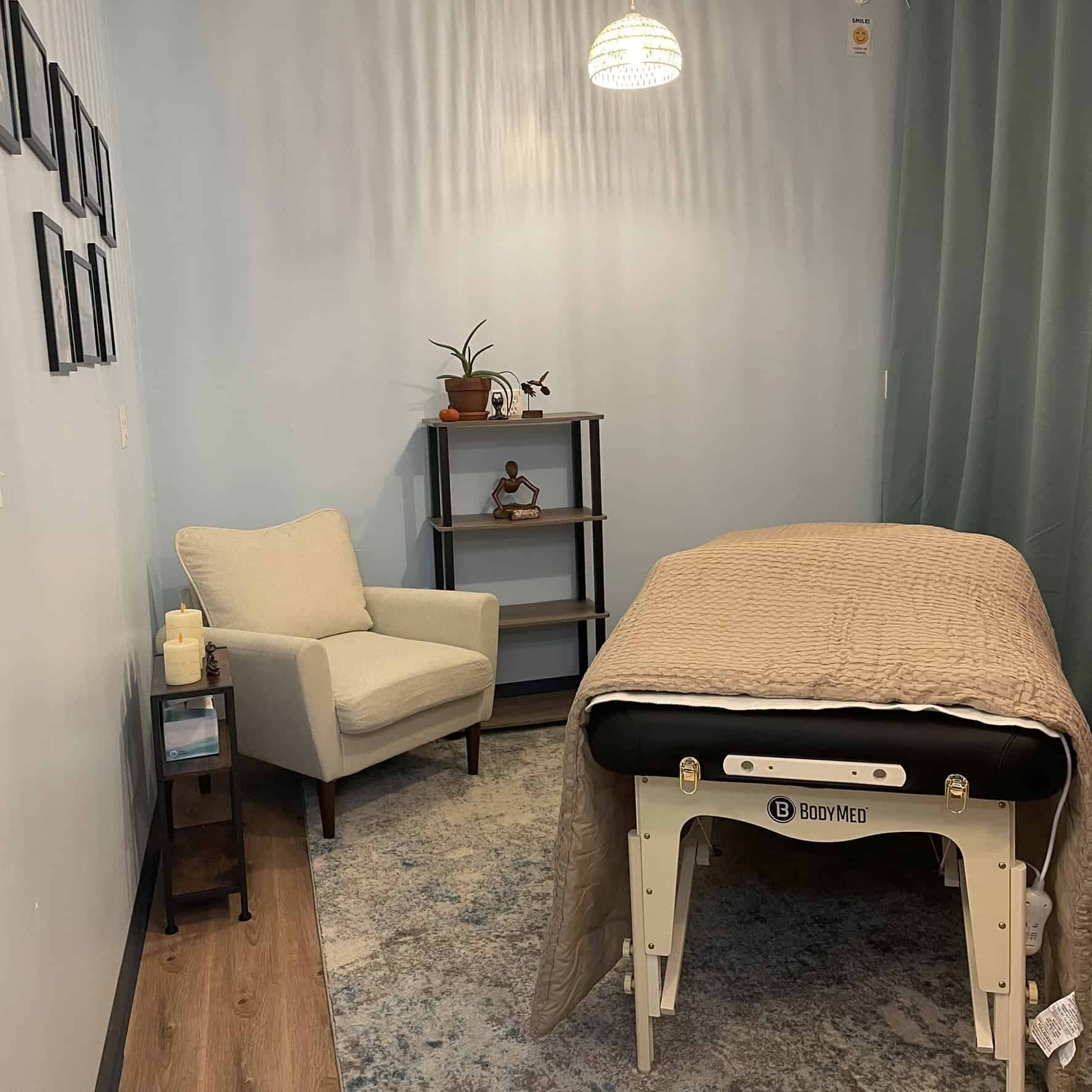Small therapy room with a massage bed covered in beige quilt, a beige chair, and a shelf with a plant, candles, and decor items against light blue walls.