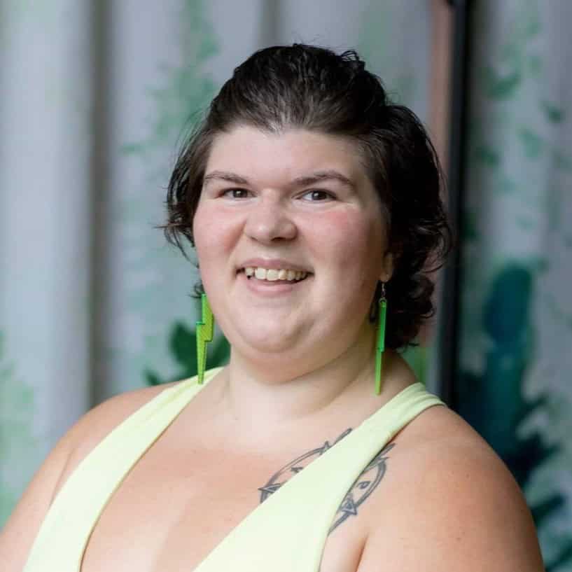 gianna Smiling woman with short dark hair, wearing a pale yellow top and bright green earrings, with a shoulder tattoo.