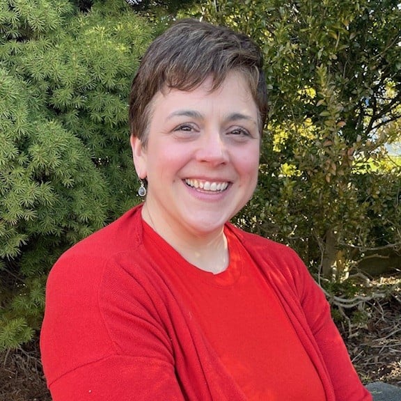 nicole-headshot Smiling woman with short hair wearing a red shirt outdoors, greenery in the background.