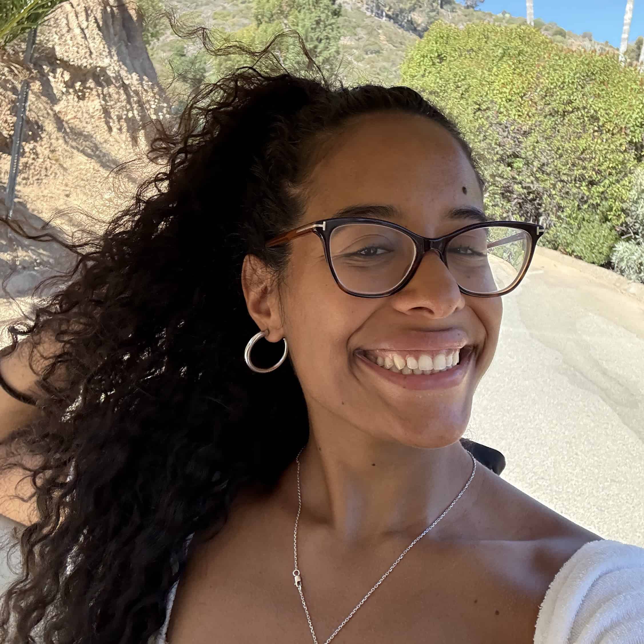 sarai-headshot Close-up selfie of a smiling woman wearing glasses and hoop earrings outdoors, with curly hair and a towel draped over her shoulder.
